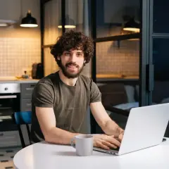 Uśmiechnięty mężczyzna z kręconymi włosami pracuje na laptopie przy kuchennym stole. Oblicza, ile wynosi czynsz za mieszkanie.