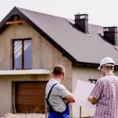 Dwóch mężczyzn omawia plany domu w budowie. Jeden z nich, w kasku, trzyma projekt. To może być kluczowe dla uzyskania kredytu deweloperskiego.
