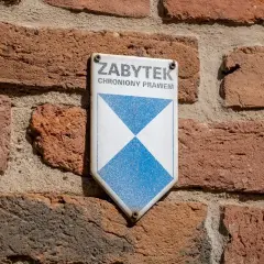 Tabliczka "ZABYTEK CHRONIONY PRAWEM" na ceglanej ścianie. To znak, że obiekt wymaga troski i konserwacji.
