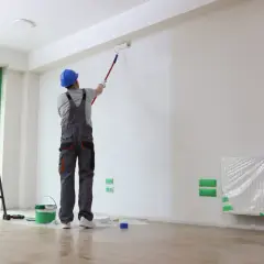 Malowanie ścian w mieszkaniu 50 m². Pracownik w niebieskim kasku i szarym ogrodniczkach maluje wałkiem białą ścianę. Obok drabina i wiadro z farbą.