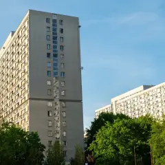 Dwa wysokie, szare bloki mieszkalne z balkonami, otoczone zielenią. W tle błękitne niebo. Taki widok to codzienność wielu lokatorów, którzy co miesiąc płacą **czynsz**.