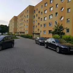 Na drodze osiedlowej zaparkowane są samochody przed blokiem z żółtą elewacją i nietypowymi oknami.