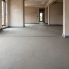Nowy, szary beton na podłodze, z niebieską taśmą przy ścianach. Widoczna dylatacja wylewki, przygotowanie do dalszych prac budowlanych.