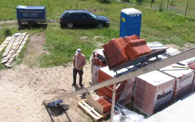 Pracownik na budowie przygotowuje dachówkę do montażu. W tle widać przyczepę, samochód i toaletę przenośną. Front robót w pełnym słońcu.