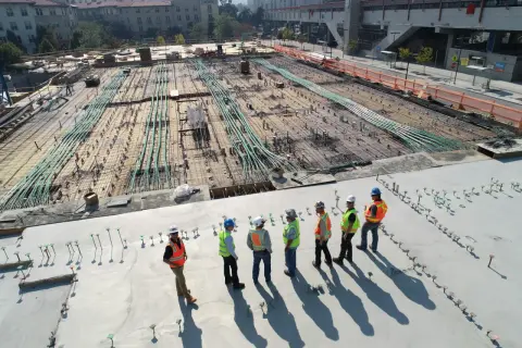 Grupa budowlańców w kamizelkach odblaskowych omawia postęp prac na budowie. W tle widać zbrojenie i instalacje, zgodne z art. 62 prawa budowlanego.