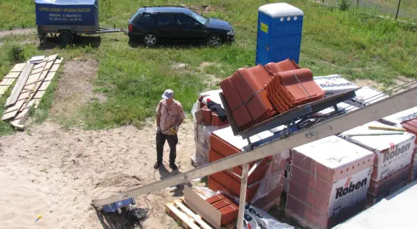 Pracownik na budowie przygotowuje dachówkę do montażu. W tle widać przyczepę, samochód i toaletę przenośną. Front robót w pełnym słońcu.