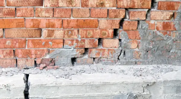 Uszkodzony mur z cegły i pęknięty betonowy cokół. Rozważasz podbijanie fundamentów? Cena może być wysoka.