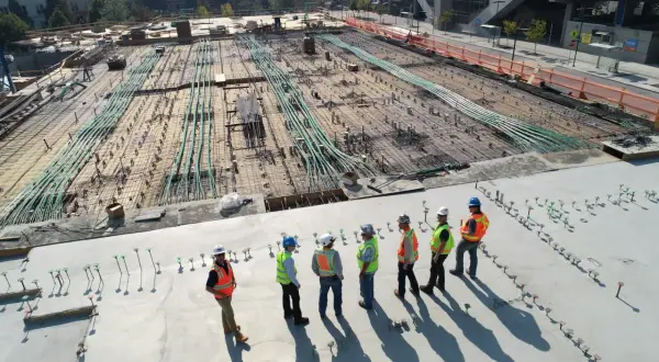 Grupa budowlańców w kamizelkach odblaskowych omawia postęp prac na budowie. W tle widać zbrojenie i instalacje, zgodne z art. 62 prawa budowlanego.