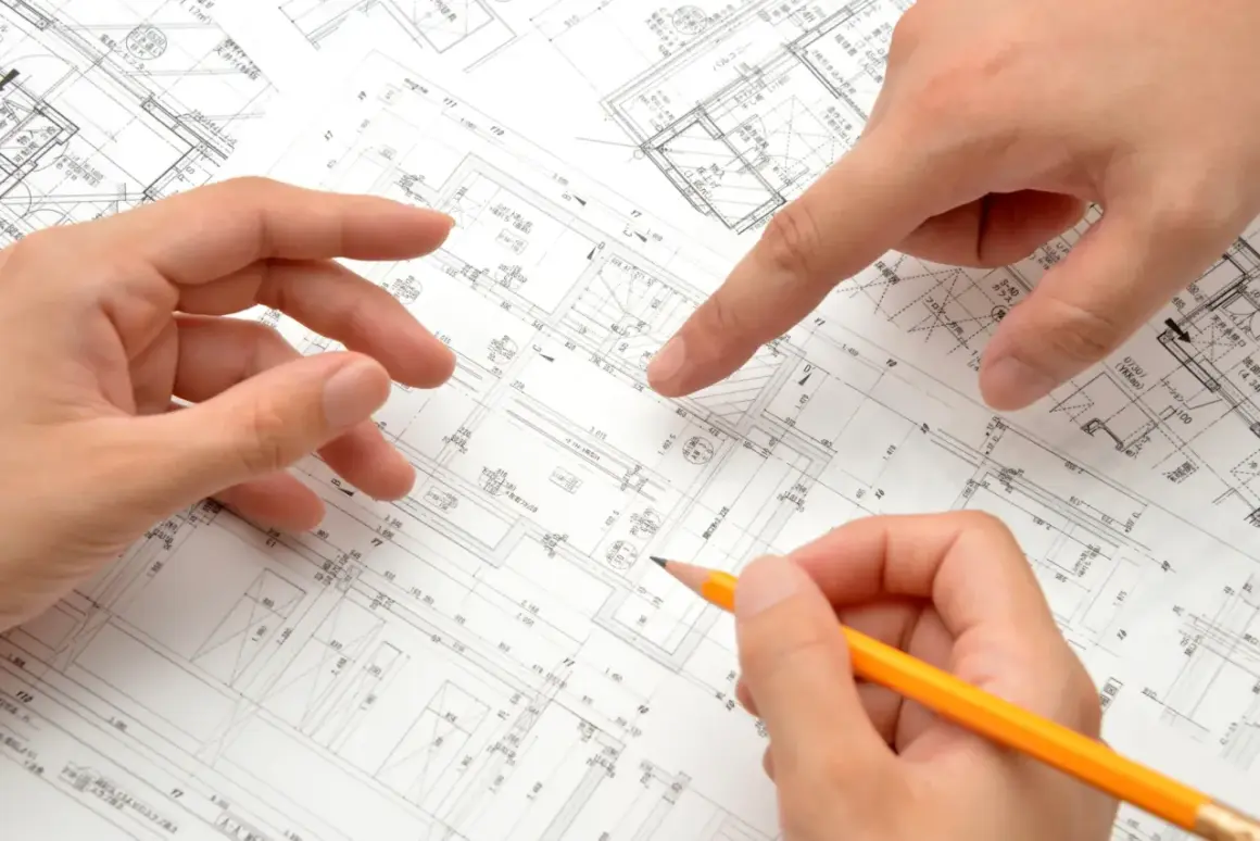 Dłonie analizują plany architektoniczne, wskazując na szczegóły dotyczące metrażu mieszkania i rozmieszczenia pomieszczeń.