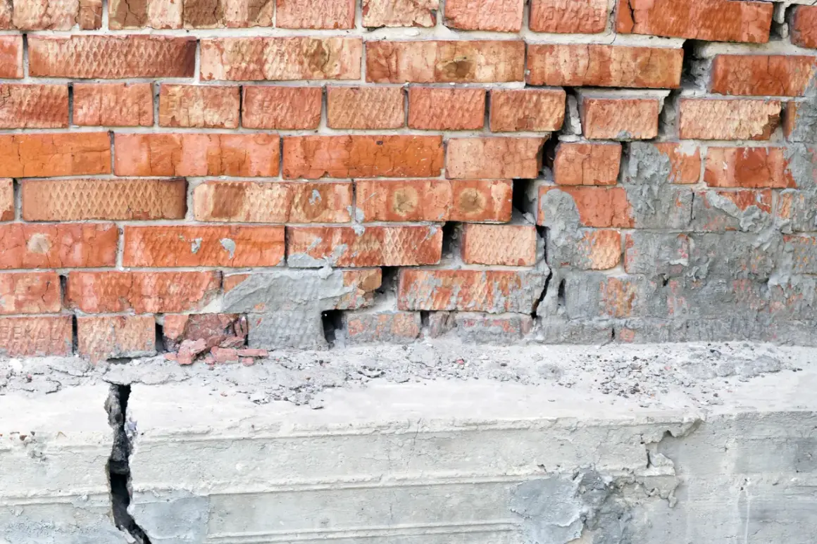 Uszkodzony mur z cegły i pęknięty betonowy cokół. Rozważasz podbijanie fundamentów? Cena może być wysoka.