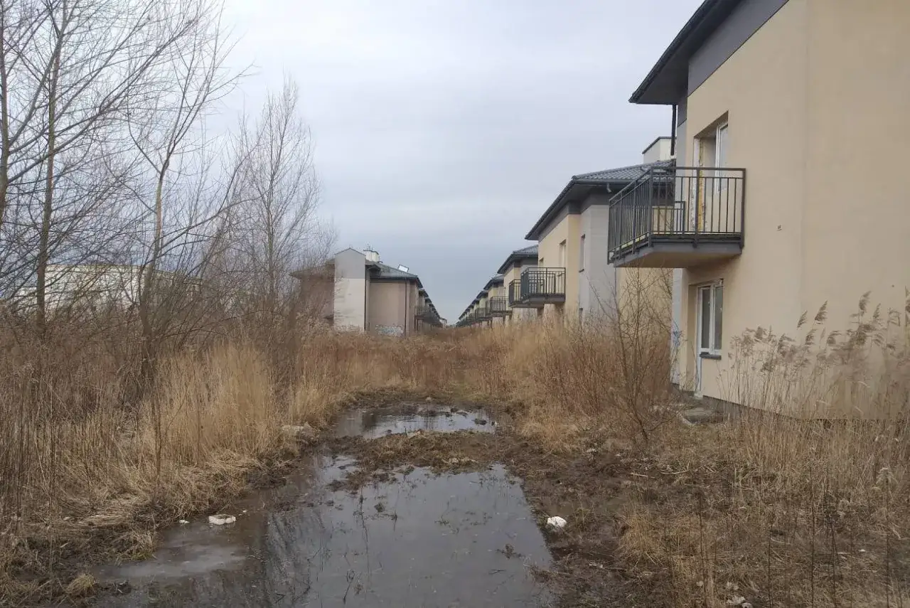 Rzędy identycznych budynków z balkonami, otoczone dziką, zarośniętą trawą i błotnistą kałużą. Obraz idealnie oddaje klimat patodeweloperki.