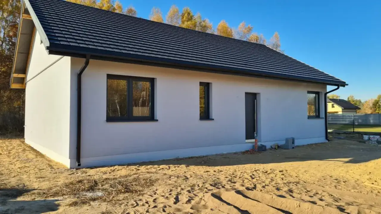 Nowy, biały dom z ciemnym dachem i oknami. Wokół piasek i drzewa. Zastanawiasz się, ile kosztuje budowa domu?