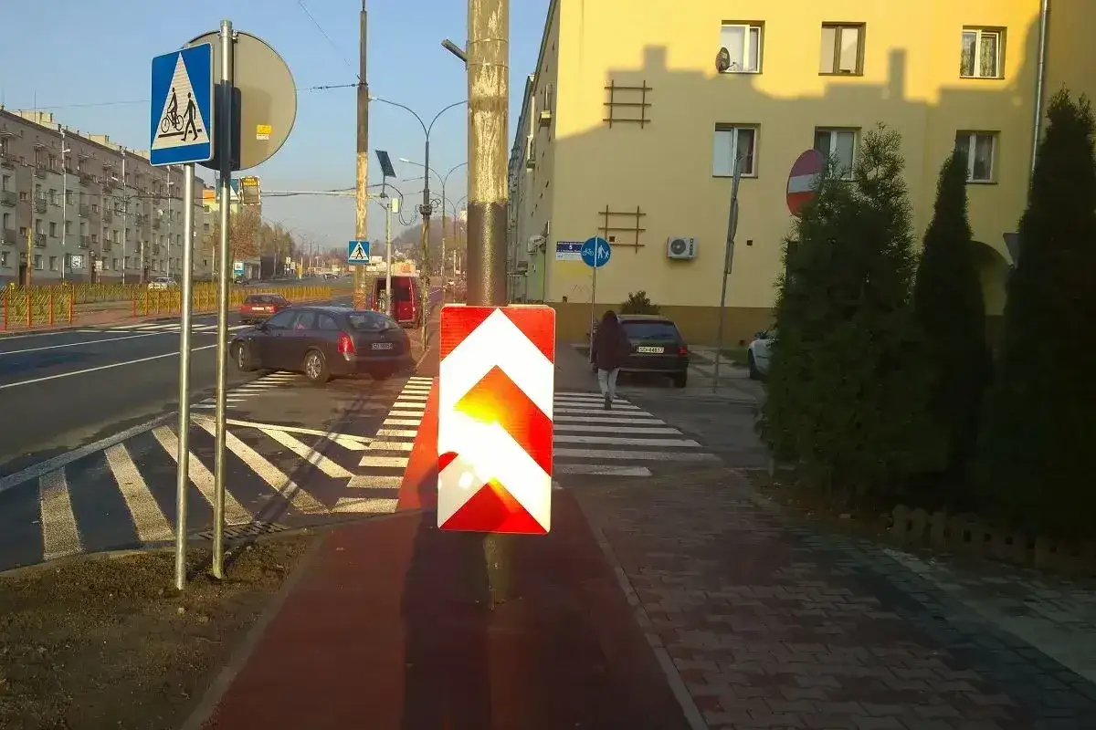 Na drodze osiedlowej znak ostrzegawczy kieruje ruch na przejście dla pieszych, gdzie osoba przechodzi przez pasy.