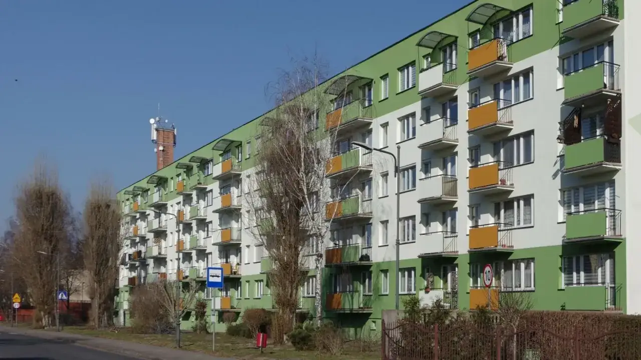 Długi blok mieszkalny z zielono-białą elewacją, balkonami i anteną na kominie. Podatek od nieruchomości mieszkanie w bloku to stały koszt.