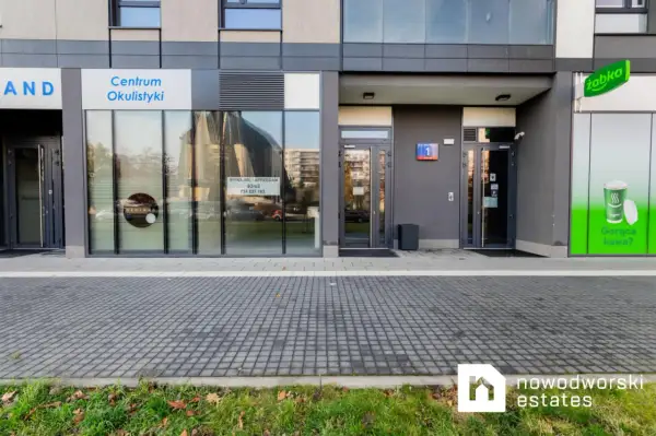 Elewacja budynku z witrynami sklepów: Centrum Okulistyki, Żabka. Widoczne szyldy i wejścia.