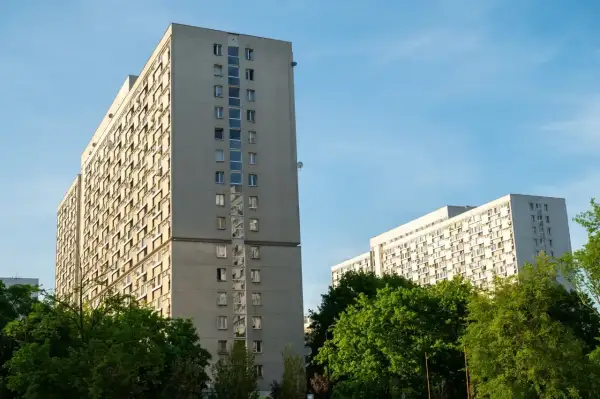 Dwa wysokie, szare bloki mieszkalne z balkonami, otoczone zielenią. W tle błękitne niebo. Taki widok to codzienność wielu lokatorów, którzy co miesiąc płacą **czynsz**.
