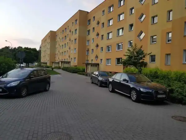 Na drodze osiedlowej zaparkowane są samochody przed blokiem z żółtą elewacją i nietypowymi oknami.